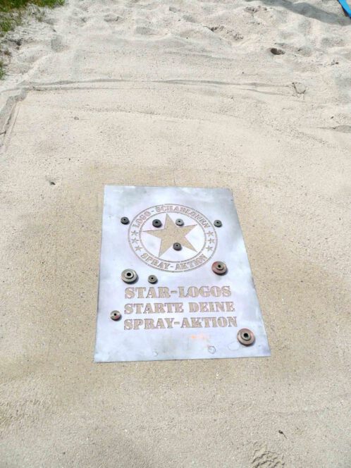 Vor der Kreidespraysignierung des Logos von Schablonen-Technik Gunnar Hansen im Sand. Schablone wurde mit Steinen beschwert. Aufnahme von vorne.