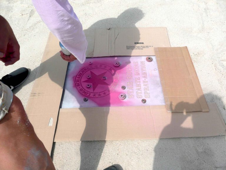 Fertige Kreidespraysignierung des Logos von Schablonen-Technik Gunnar Hansen im Sand. Farbe pink. Aufnahme von der Seite, mit menschlichem Schatten