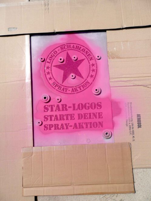 Fertige Kreidespraysignierung des Logos von Schablonen-Technik Gunnar Hansen im Sand. Farbe pink. Aufnahme von vorne.