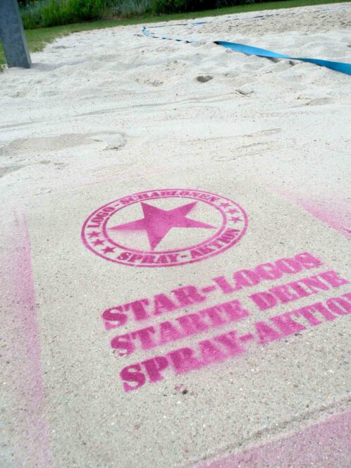 Fertige Kreidespraysignierung des Logos von Schablonen-Technik Gunnar Hansen im Sand. Farbe pink. Aufnahme von schräg vorne.