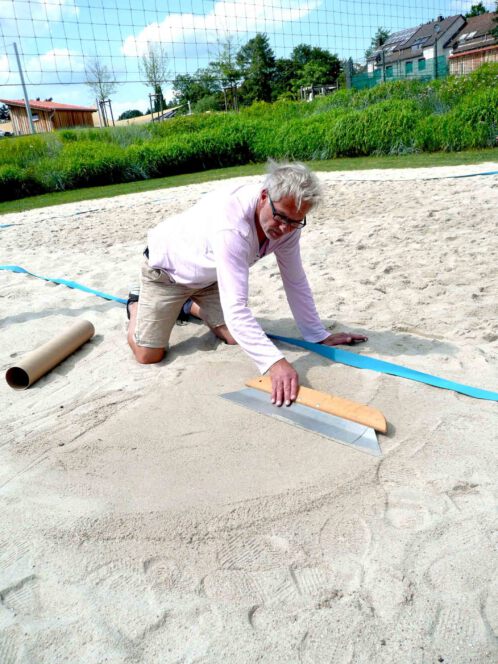 Gunnar Hansen beim Glätten einer Sand- und Strandfläche mit breiter Flächenspachtel zur Vorbereitung für Sprühaktion mit Schablone und Kreidespray.
