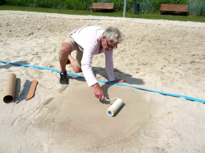 Gunnar Hansen beim Glätten einer Sand- und Strandfläche mit breiter Andruckrolle zur Vorbereitung für Sprühaktion mit Schablone und Kreidespray.