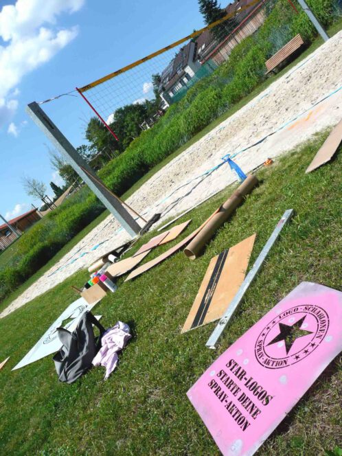 Logo- und Sloganschablone von Schablonen-Technik Gunnar Hansen für Kreideaktion und Fotoshootings liegen im Gras verteilt. Zu sehen sind auch Werkzeuge zur Herstellung temporärer Kennzeichnungen auf Sand.