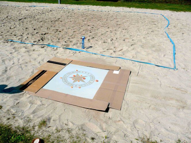 Logoschablone von Schablonen-Technik Gunnar Hansen liegt flach im Sand eines Volleyballfelds. Vorbereitung für eine Sprühaktion mit Kreidespray. Ränder mit Pappstreifen abgedeckt. Das dient zum Schutz vor Sprühnebel