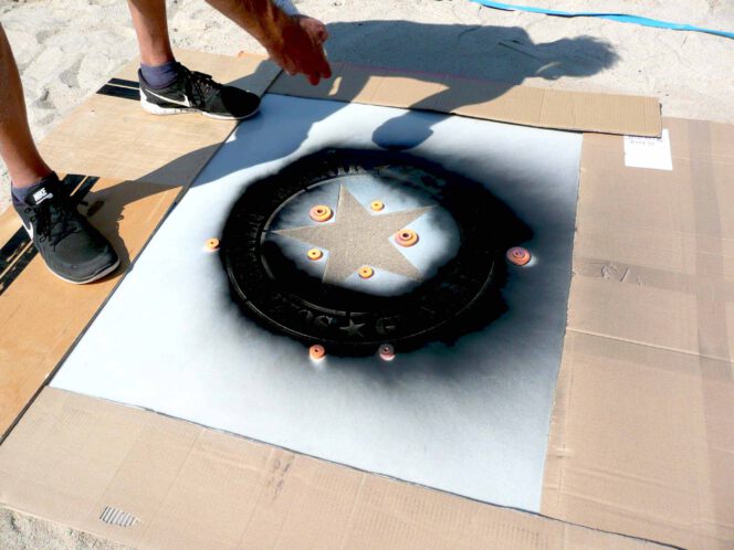 Sprühen des Logos von Schablonen-Technik Gunnar Hansen auf einem Volleyballfeld mit Kreidespray. Farbe schwarz. Ränder mit Pappstreifen abgedeckt. Das dient zum Schutz vor Sprühnebel