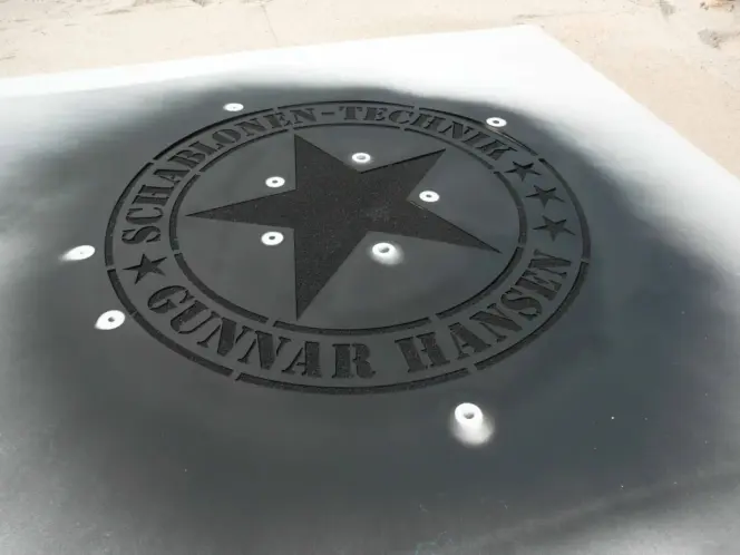 Fertige Kreidespraysignierung des Logos von Schablonen-Technik Gunnar Hansen im Sand. Farbe schwarz. Die Schablone liegt noch im Sand.