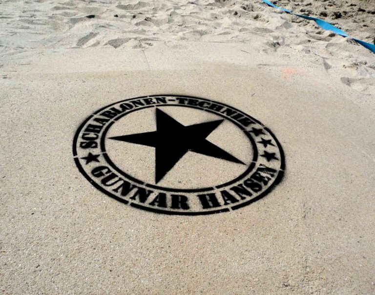 Fertige Kreidespraysignierung des Logos von Schablonen-Technik Gunnar Hansen im Sand. Farbe schwarz. Aufnahme von schräg vorne.