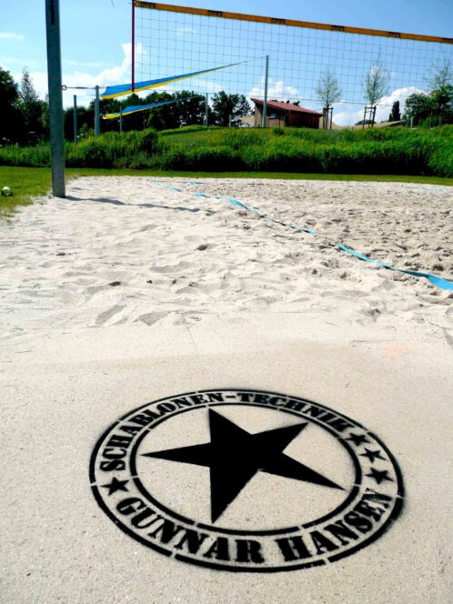 Fertige Kreidespraysignierung des Logos von Schablonen-Technik Gunnar Hansen auf einem Volleyballfeld. Farbe schwarz. Aufnahme von schräg vorne. Im Hintergrund Gras und Sonnensegel