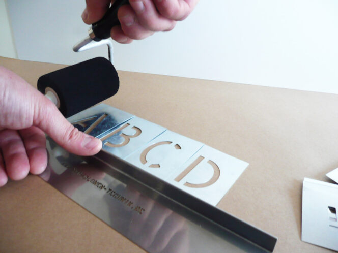 Die Stencil-Schiene mit den Buchstaben-Schablonen ABCD wird mit der linken Hand auf Karton gedrückt, in mit der Rechten Hand wird der Signierroller über die Schablone gehalten..