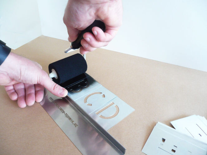 Die Stencil-Schiene mit den Buchstaben-Schablonen ABCD wird mit der linken Hand auf Karton gedrückt, in mit der Rechten Hand wird der Signierroller geführt.